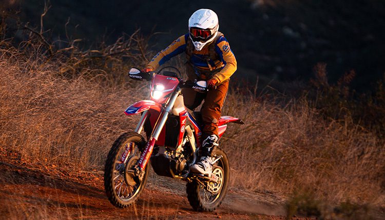 Red Motorcycle with rider at dusk in the dirt with light on.