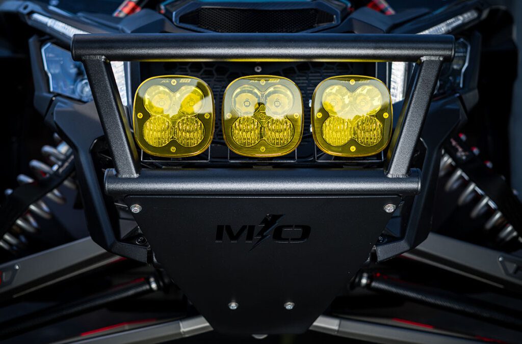 Close-up of a Baja Designs off-road lighting setup on a Can-Am Maverick X3, featuring three yellow-lens LED lights mounted on a rugged MLO front bumper. This high-performance UTV lighting system enhances visibility for extreme off-road adventures and racing conditions.