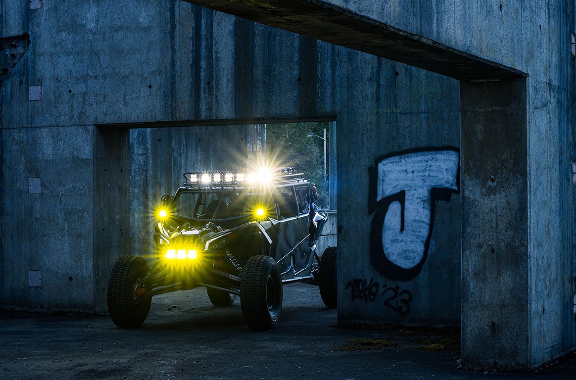 Off-road Can-Am Maverick X3 with powerful Baja Designs lighting setup, illuminating a rugged urban environment with bright yellow and white lights. The vehicle is positioned under a concrete structure with graffiti in the background, highlighting its aggressive and adventurous design.